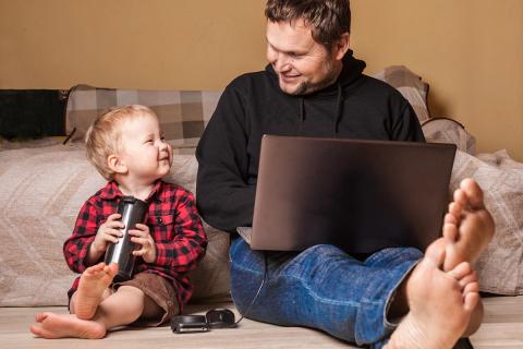 Father works on laptop as young son happily sits next to him.