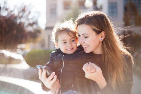 Mother holds child's hands as she looks at her phone outside on a crisp Autumn day.