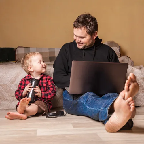 Father works on laptop as young son happily sits next to him.