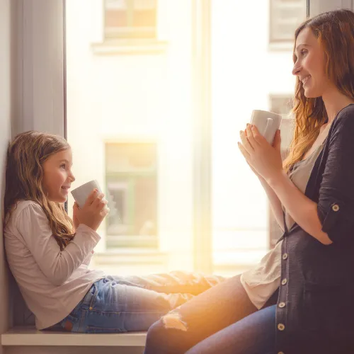 Mother and daughter sit in windowsill while holding mugs
