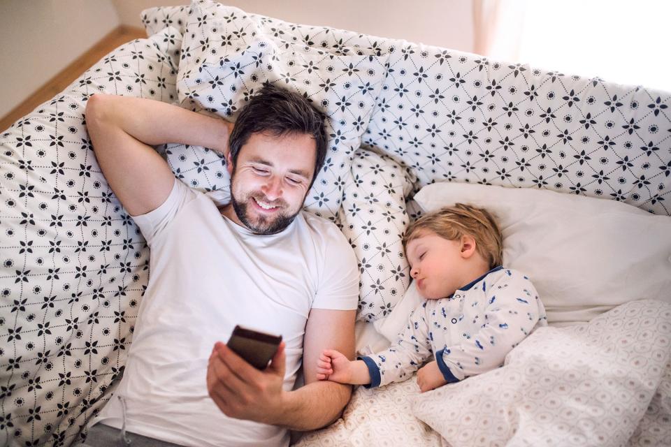 Father laughs while looking at his phone while reclining on the couch next to his sleeping toddler.