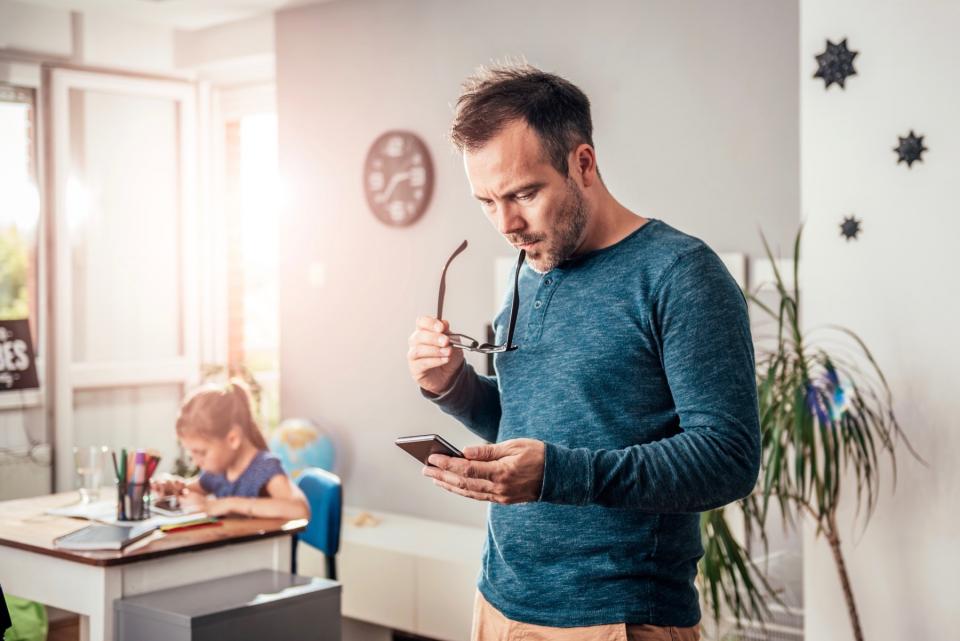 A man stands and looks at his phone while his daughter draws at the table.