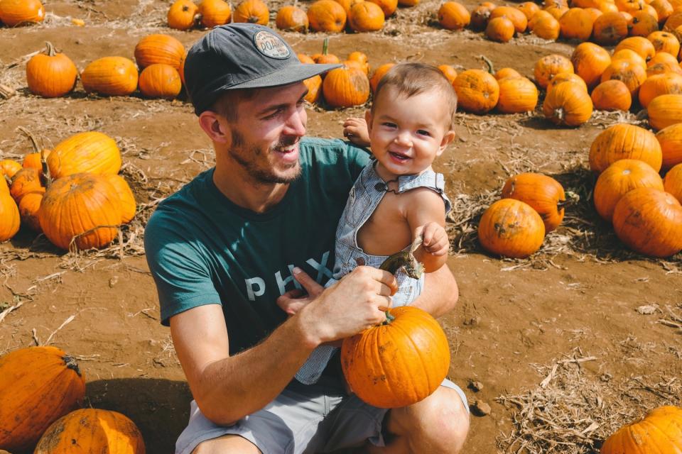 Father and infant son smile together in a pumpkin patch.