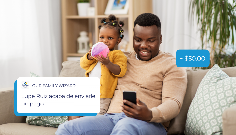 Una niña está de pie en el sofá junto a su padre mientras él mira a su teléfono para verificar el reembolso de los gastos.